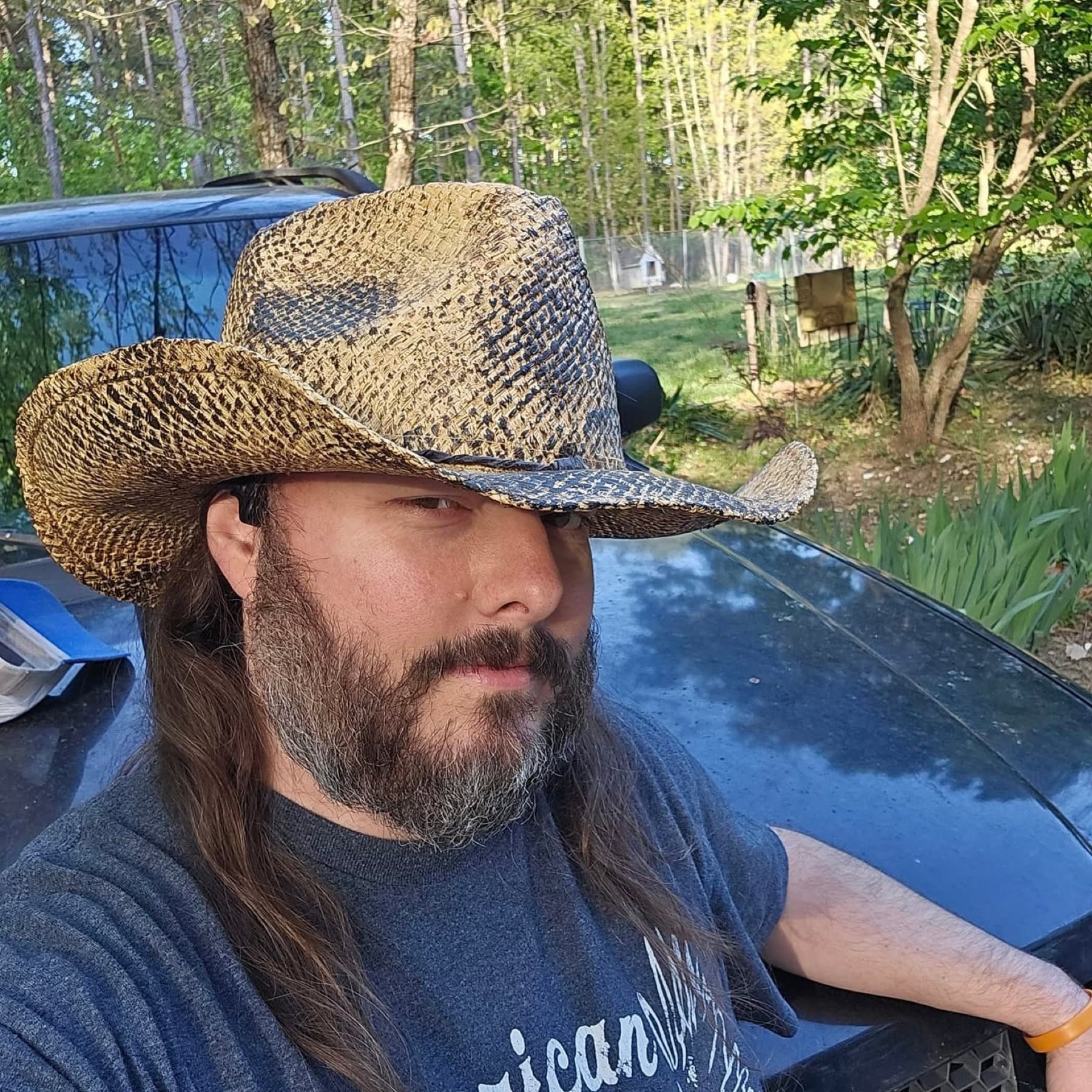 Recent portrait of Jawa (Shawn, @JawaMeta) wearing a woven straw cowboy hat, long hair and beard, leaning on a dark truck